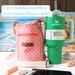 An orange bag with a circle print, is pictured beside a Stanley 40 ounce Quencher, chap stick, money and a cell phone.
